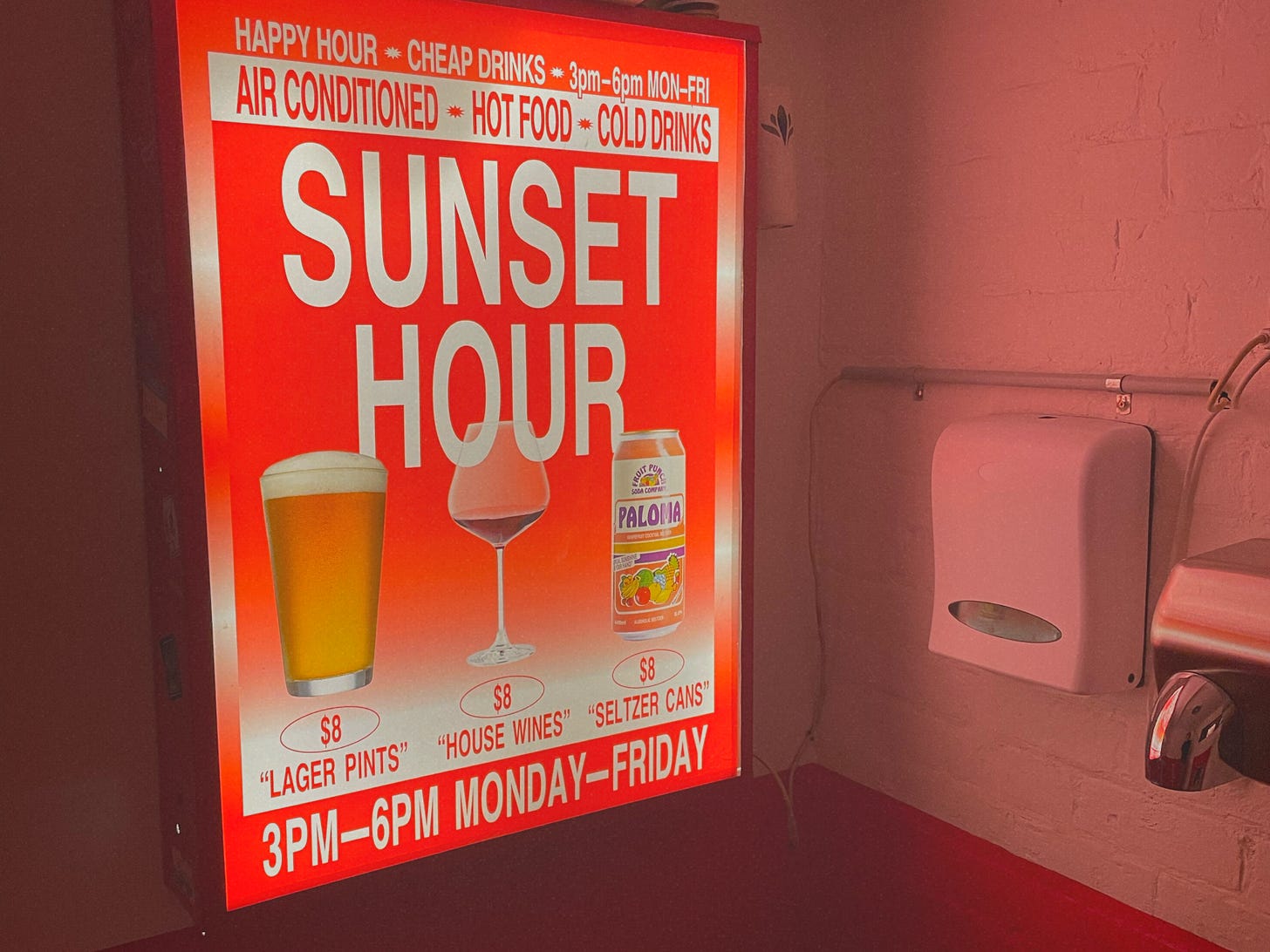 illuminated advert for "Sunset Hour" drinks on a screen in a public bathroom