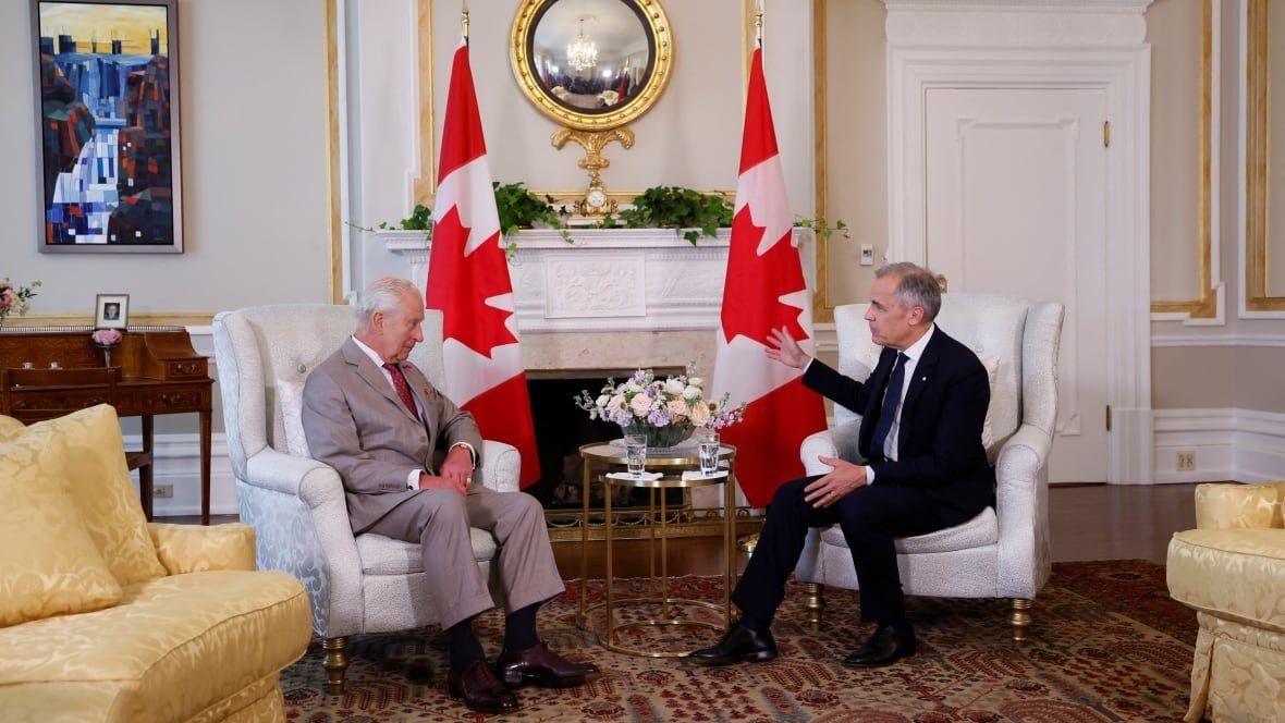 Two men in suits sit at silver chairs in front of a fireplace.
