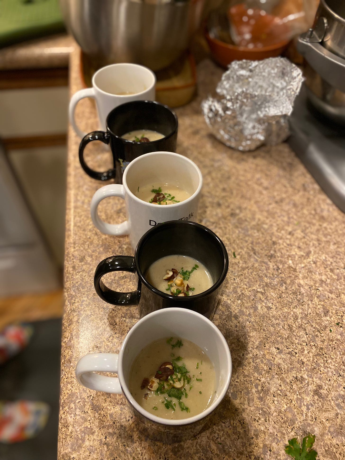 Five mugs of creamy soup topped with herbs and toasted hazelnuts, sitting on a counter somewhat cluttered in the background.