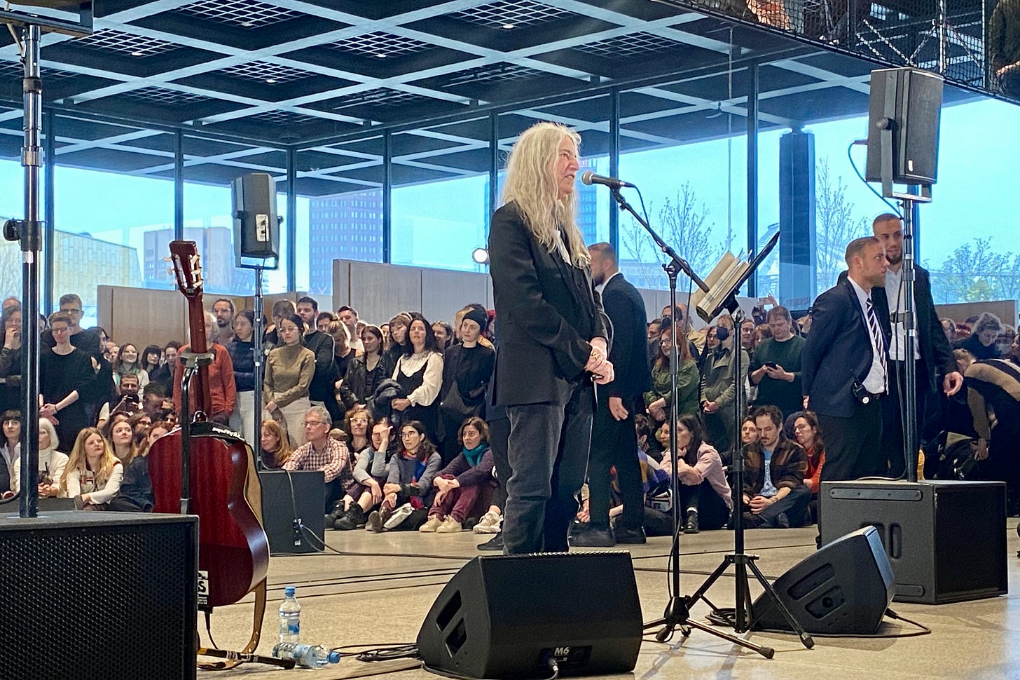 A imagem mostra a artista Patti Smith em pé, vestindo um terno preto. Ela está em frente a uma plateia sentada no chão. Ao fundo, podemos ver uma parede de vidro que reflete o público.