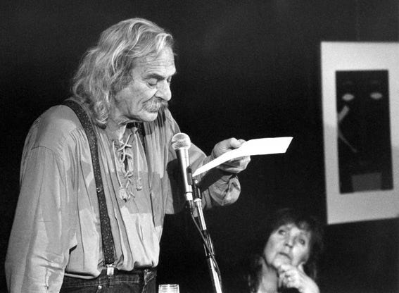 A black and white photo of Jack Hirschman standing at a microphone reading from a piece of paper.