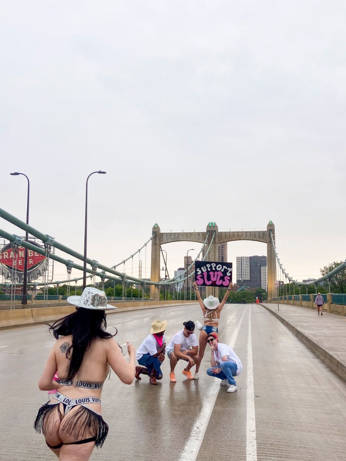 a woman in short shorts and a white cowboy hat holds up a sign reading "support sluts" while three men in white tshirts kneel behind her on the a bridge, a girl in a disco cowboy hat walks toward them