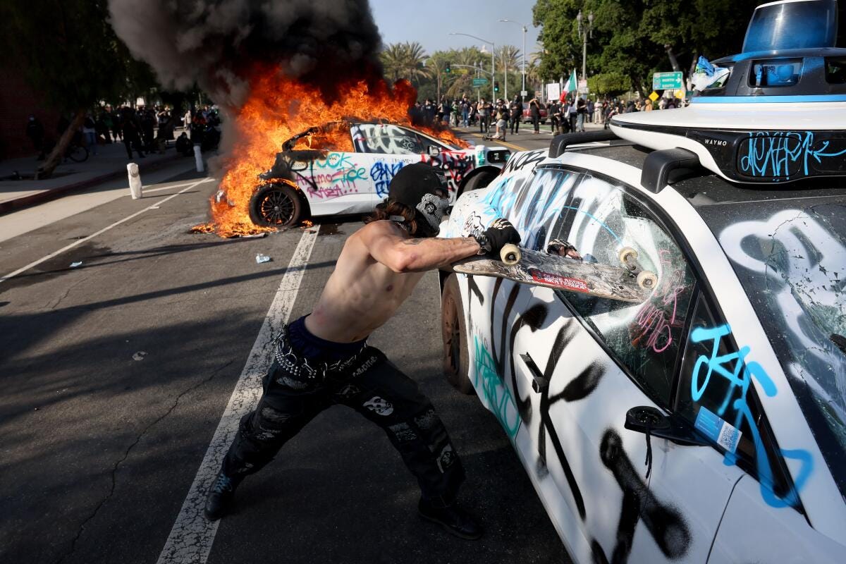 A person damages a vehicle while another vehicle burns in the background.