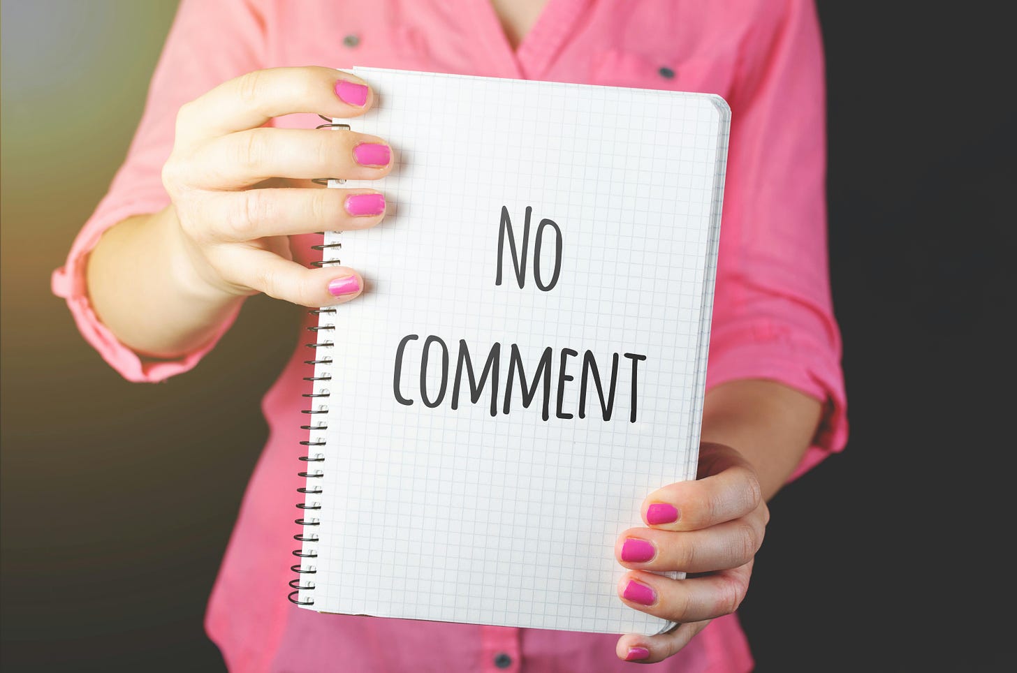 A person in a pink button up shirt, with pink nails, holds up a noteboo with the word “no Comment” written on it in black ink