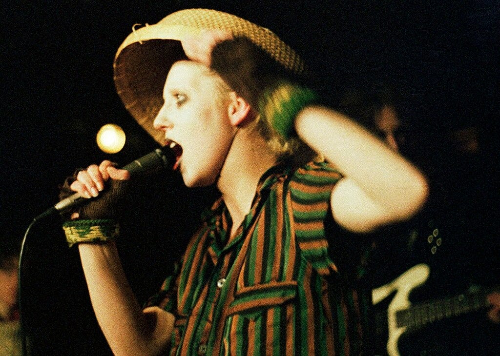 Close up of Jayne singing into a microphone. She's wearing a striped shirt and has short hair under a straw hat.