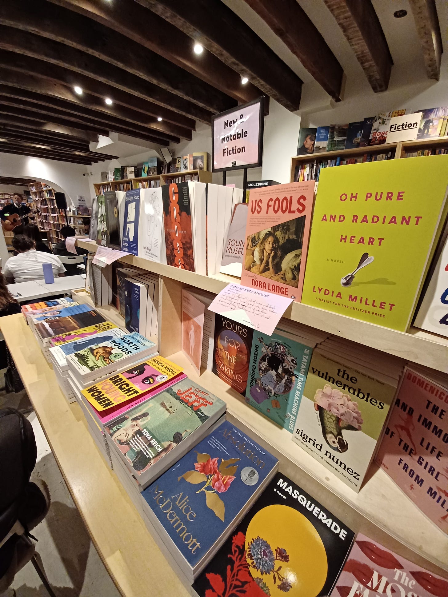 A picture of two shelves within Books Are Magic, a bookstore in Brooklyn, NY. The shelf says "New and Notable Fiction," and has a display of various, colorful paperback books in two rows, where the books rest upright, and under the shelf are books laying flat.