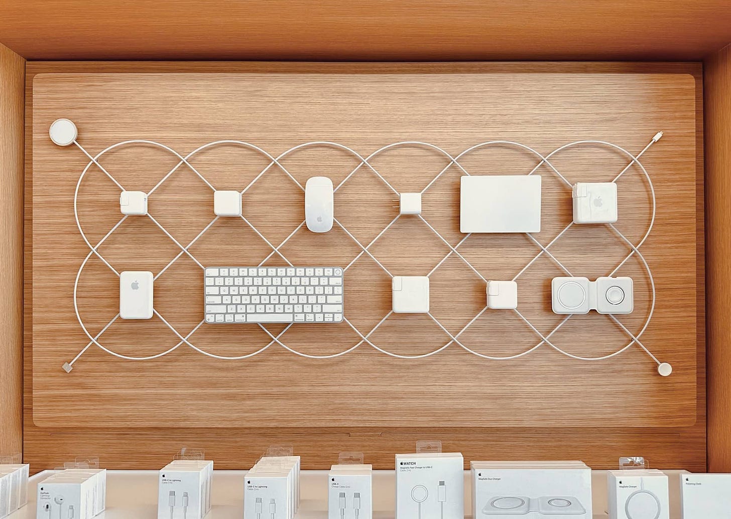 The new Essentials bay at Apple Park Visitor Center. iPhone, iPad, and Mac accessories are represented on a wood background with braided MagSafe chargers weaving a pattern in the background.
