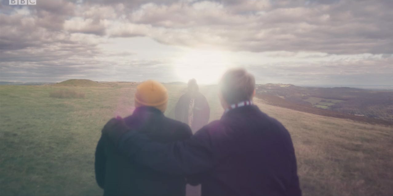 This image shows Ryan Sinclair (played by Tosin Cole, left) and Graham O'Brien (played by Bradley Walsh, right) standing in a Sheffield, England field and see a sunlit vision of Grace Sinclair-O'Brien (played by Sharon D. Clarke) in the final moments of "Revolution of the Daleks" (Doctor Who's 2021 New Year's Day Special). Ryan and Graham stand with their backs to camera, while Grace faces them from several feet away. Graham comforts Ryan by holding Ryan's left shoulder with his (Graham's) left arm. This image shows Ryan Sinclair (played by Tosin Cole, left) and Graham O'Brien (played by Bradley Walsh, right) standing in a Sheffield, England field and see a sunlit vision of Grace Sinclair-O'Brien (played by Sharon D. Clarke) in the final moments of "Revolution of the Daleks" (Doctor Who's 2021 New Year's Day Special). Ryan and Graham stand with their backs to camera, while Grace faces them from several feet away. Graham comforts Ryan by holding Ryan's left shoulder with his (Graham's) left arm.