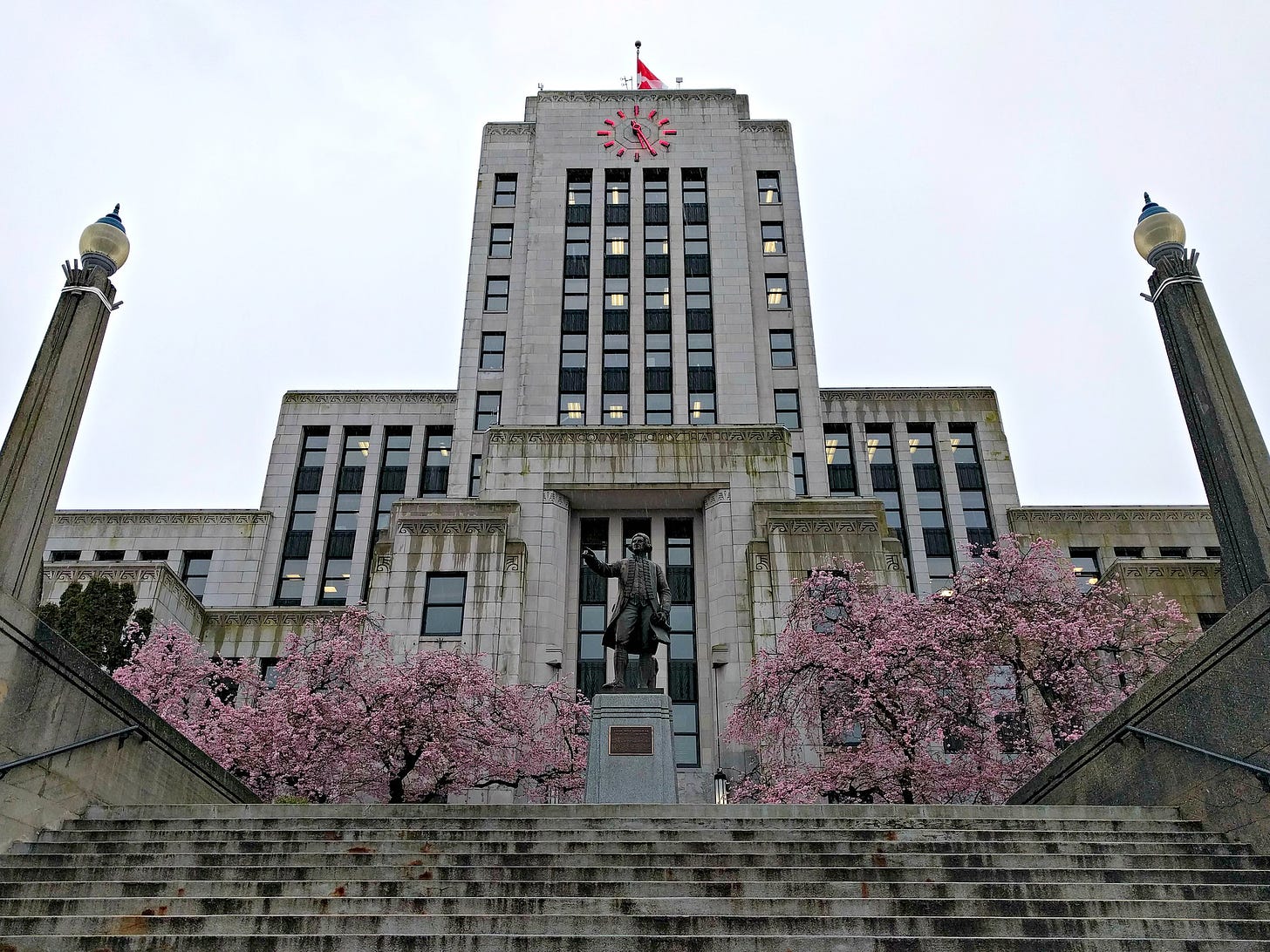 Vancouver City Hall looms in front of the viewer. The shot is taken from the bottom of the stairs in front of city hall, with the statue of, I dunno, some guy in front. Cherry blossoms are blooming around the statue and entrance to city hall