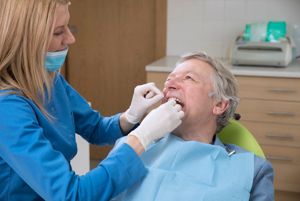 A dental professional examines an older man's teeth in a dental clinic. They both wear protective gear.