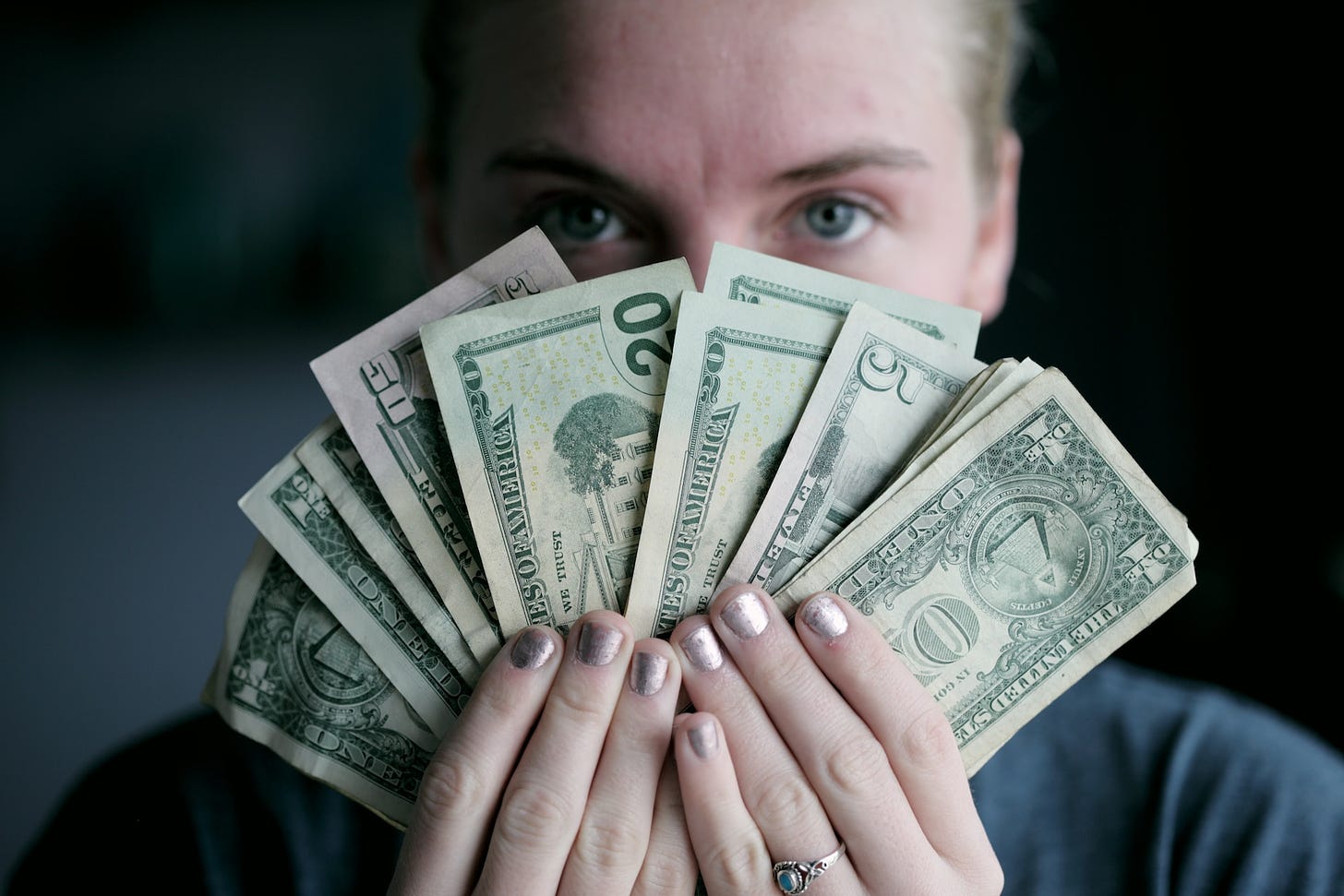 Photo of a woman's face, just showing her eyes and forehead. In front of her face, she is holding American money with both hands, in a fan shape of different denominations.
