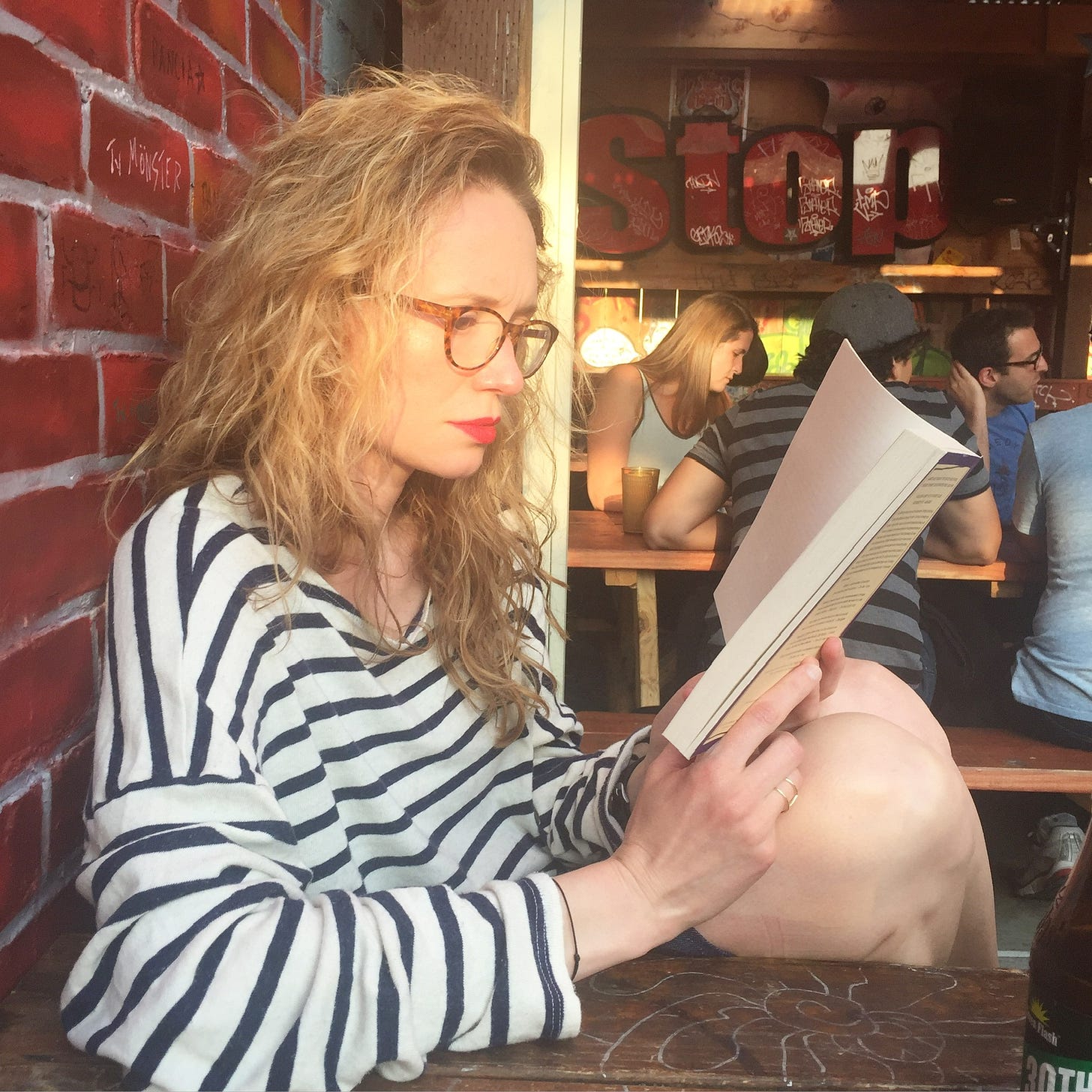 A white woman with glasses and long hair reads a book at a bar