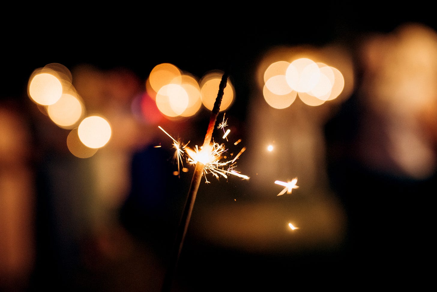 An image of a sparkler aflame, with about half of the sparkler left to burn. In the background, blurry lights, yellow and red, and indeterminate shapes. 