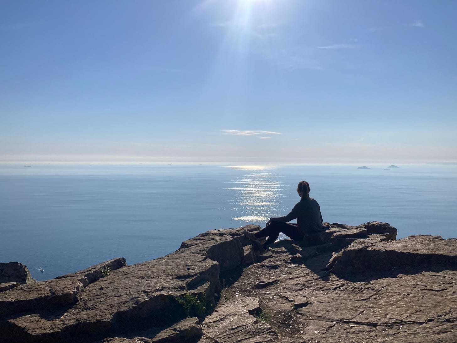 Girl sitting on top of rocky ledge looking out to sea.