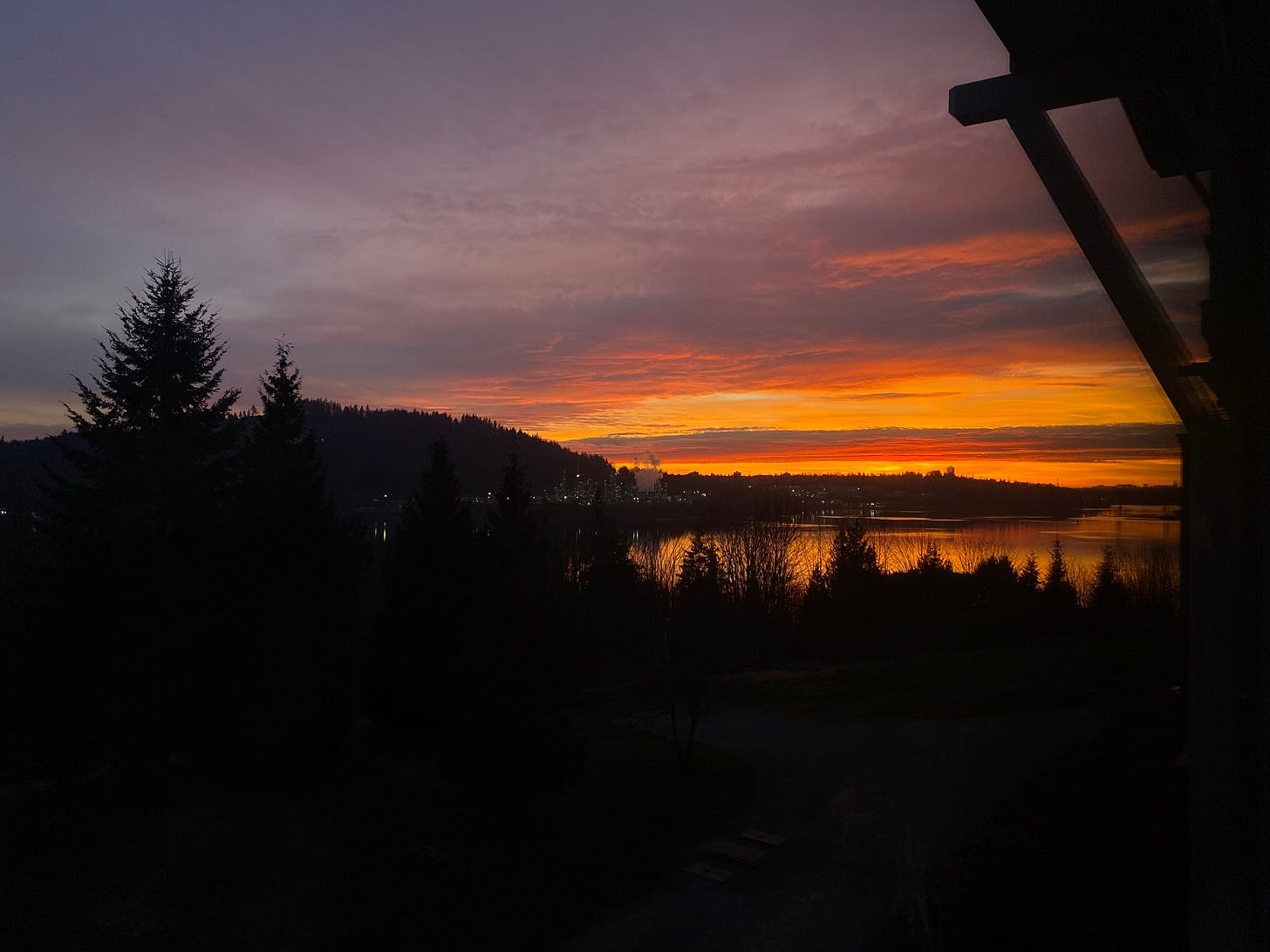 The inlet at sunset. The trees and landscape in the foreground are all in silhouette, as is Capitol Hill across the water. The sky is a bluish mauve just above the hill, with vibrant oranges and magenta at the horizon. The colours of the sunset are reflected on the ocean.