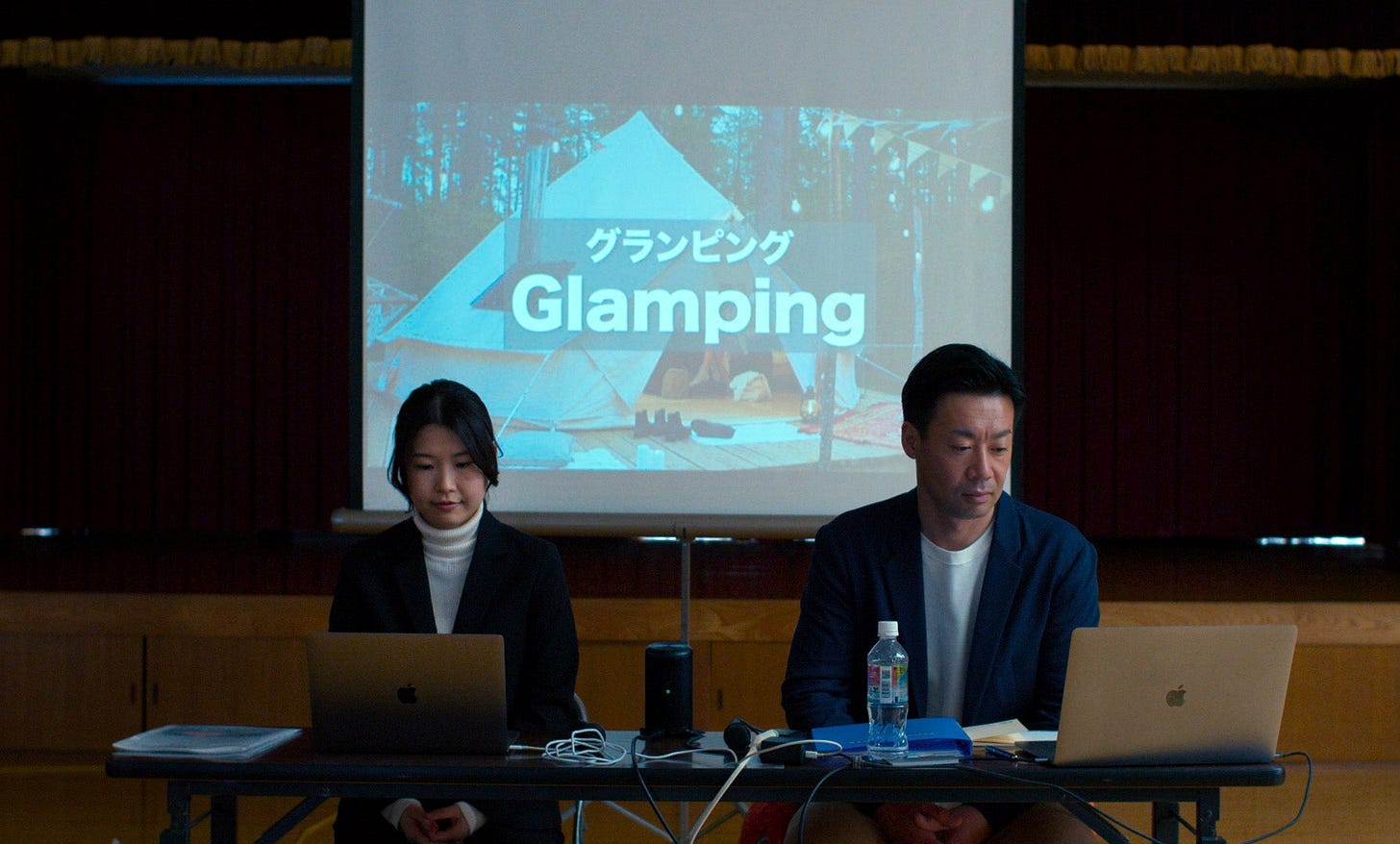 A woman (Ayaka Shibutani) and a man (Ryuji Kosaka) wearing suits and sitting in front of a powerpoint slide that says 'Glamping'