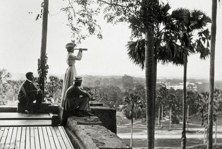 Victorian era black and white photo of a woman looking out over a palm grove with a spyglass.