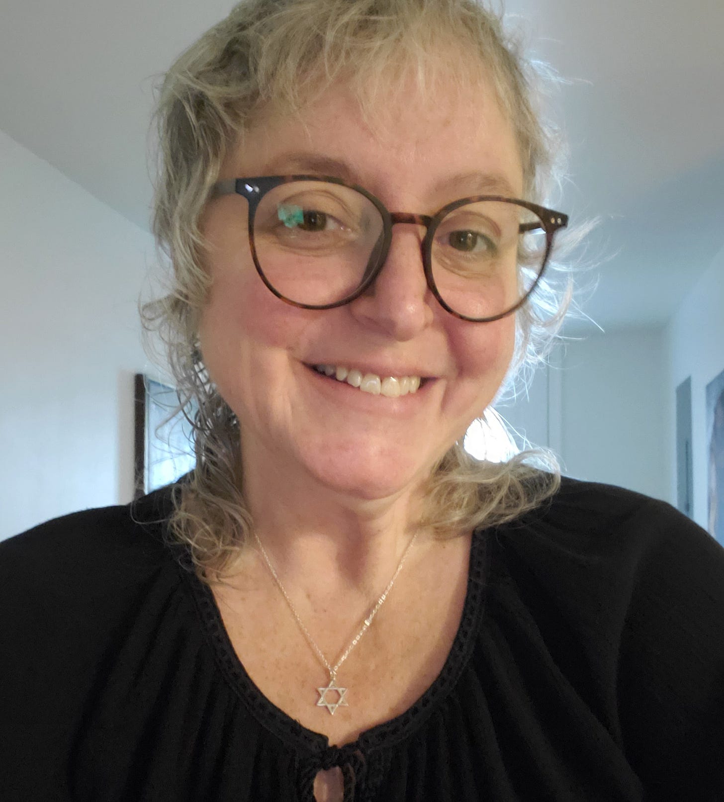 A white woman with gray hair and round glasses sporting a Jewish star necklace. 