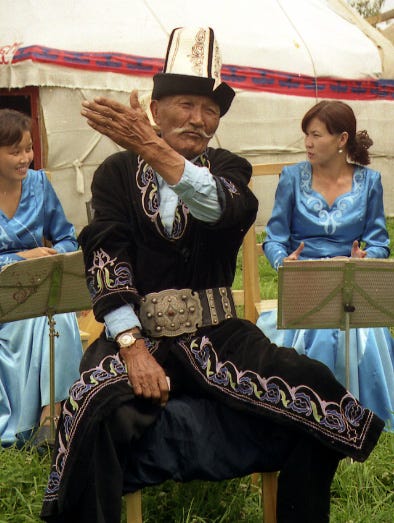 An older man gestures during a storytelling event. He is wearing a high crowned white hat with a felt lined brim. Women in blue are in the background