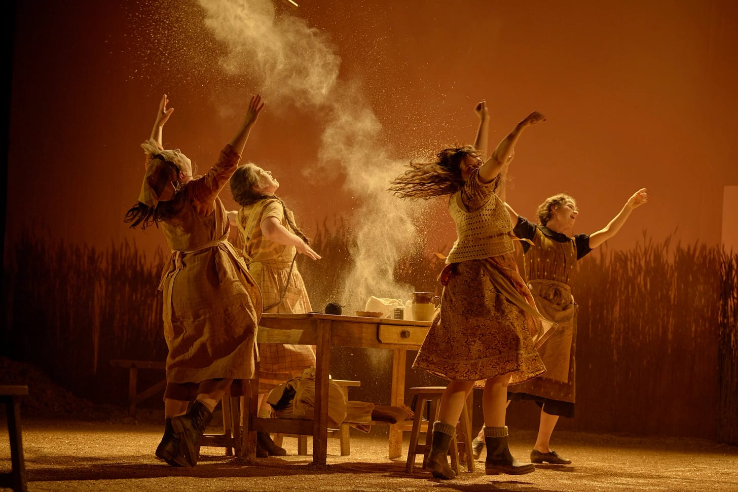 Four women wearing dresses and aprons are standing up around a wooden kitchen table. Their arms are thrown up into the air, as their bodies are frozen into a still of motion. One of the women has thrown flour in the air, where it hangs to capture the ecstasy of the moment. 