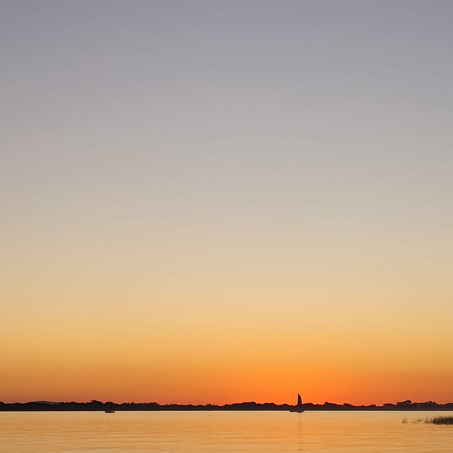 imagem do horizonte de um rio, ao por do sol. o céu possui tons entre azul claro e alaranjado. há um barquinho flutuando ao longe.