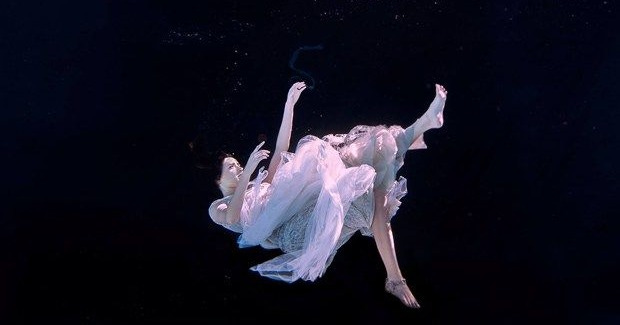 This may contain: a woman floating in the water wearing a white dress and holding her leg up with both hands