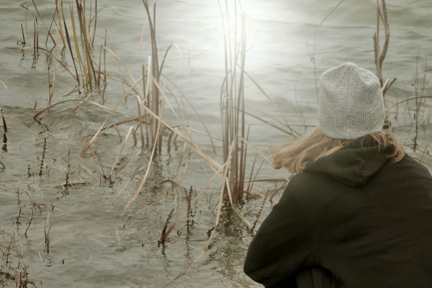 A young woman in a gray knit cap with long blond hair looks sits on the bank looking out across the shore with lake grass in the forefront and the sun light shining in the muted colors of the lake