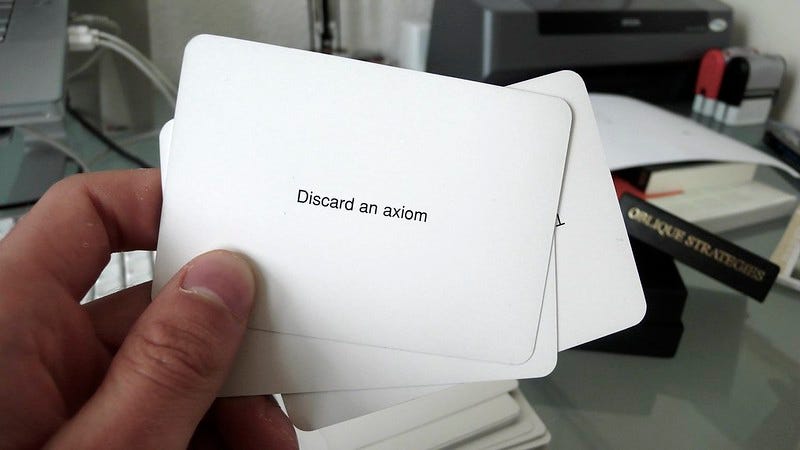 Close-up photograph of a hand holding several cards from the Oblique Strategies deck, desk in background.