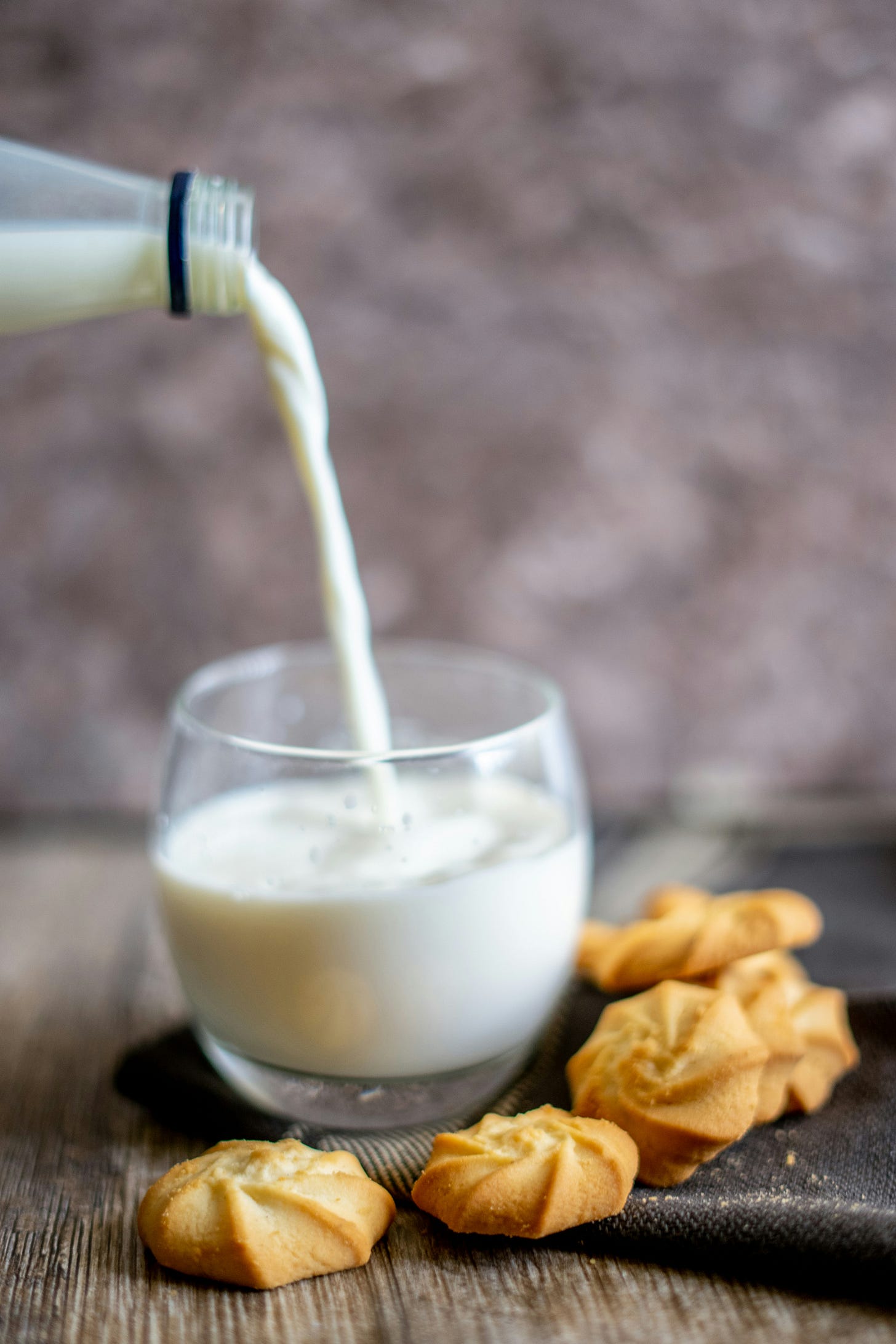 Milk is poured from a bottle into a round glass beaker. Next to the cup is a pile of little buttery cookie swirls.