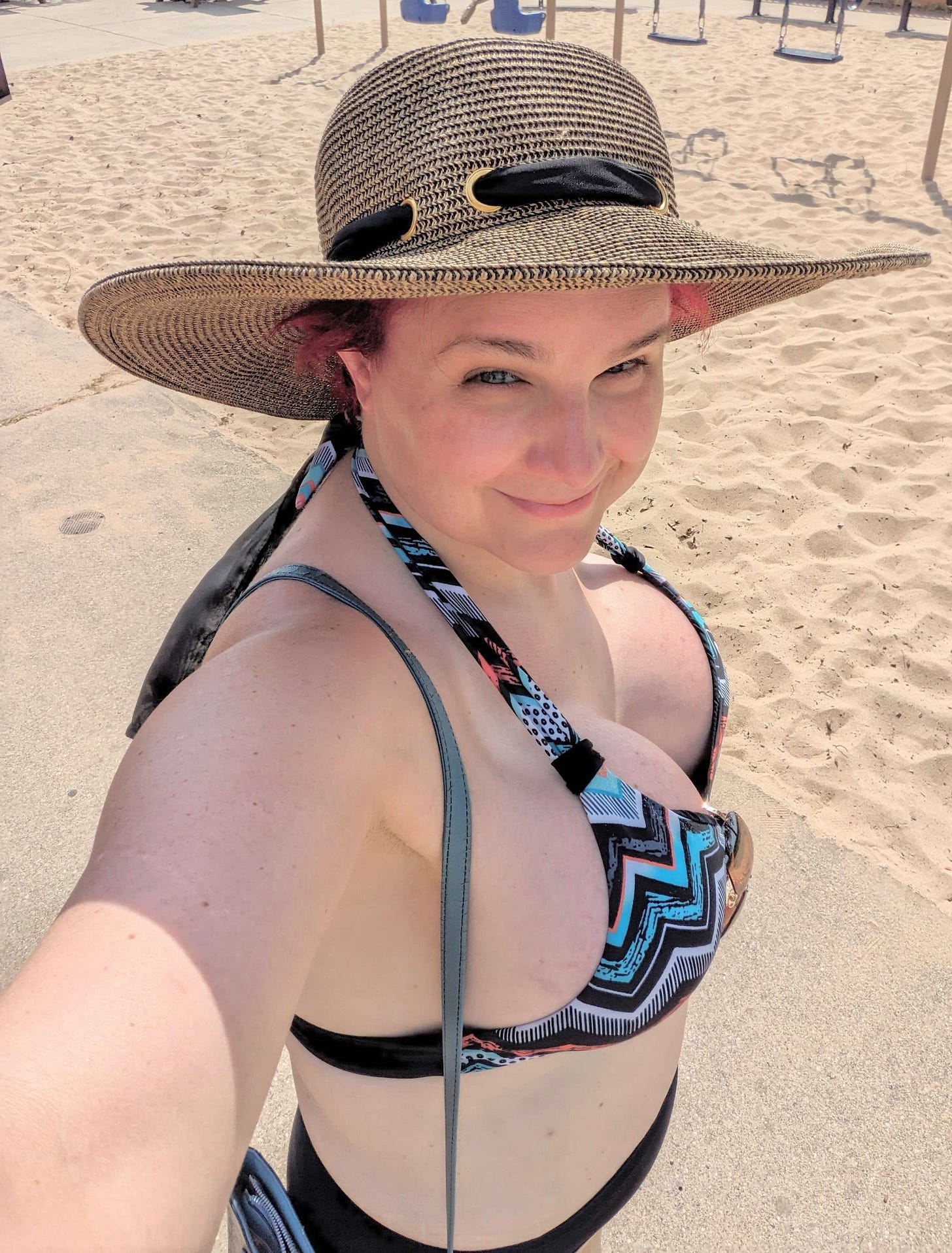 The author, at the beach, wearing a black and green halter-style bikini and a sun hat with a black ribbon around the brim.