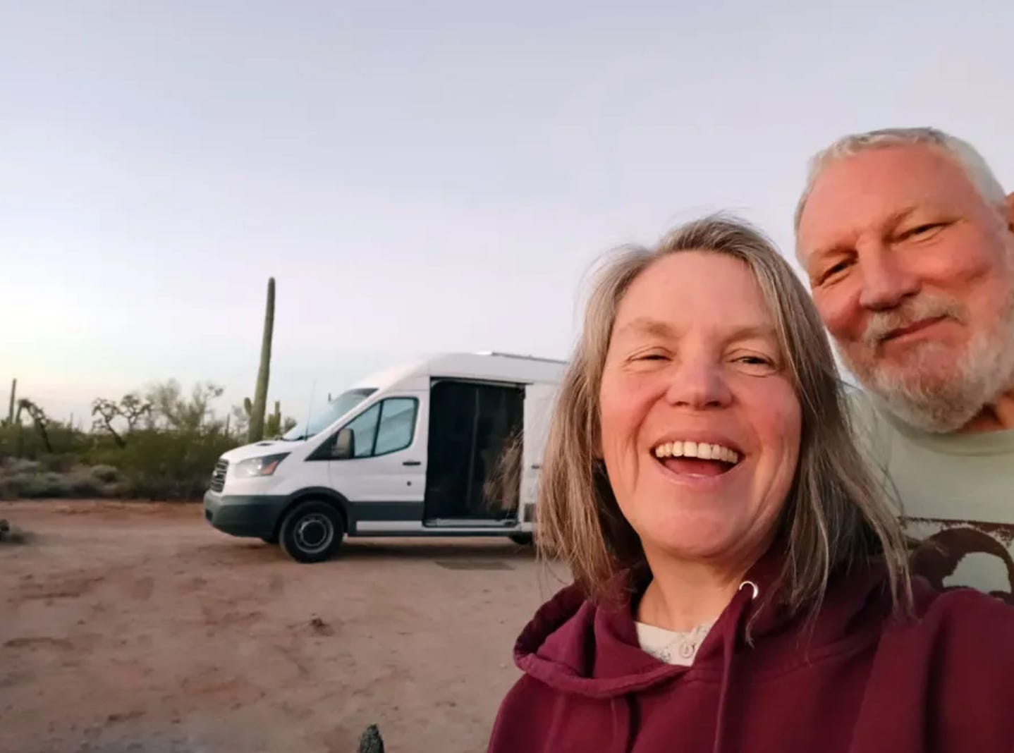 A woman and a man take a selfie in a desert, behind them a white van they call Ballena Blanca, their home on wheels.