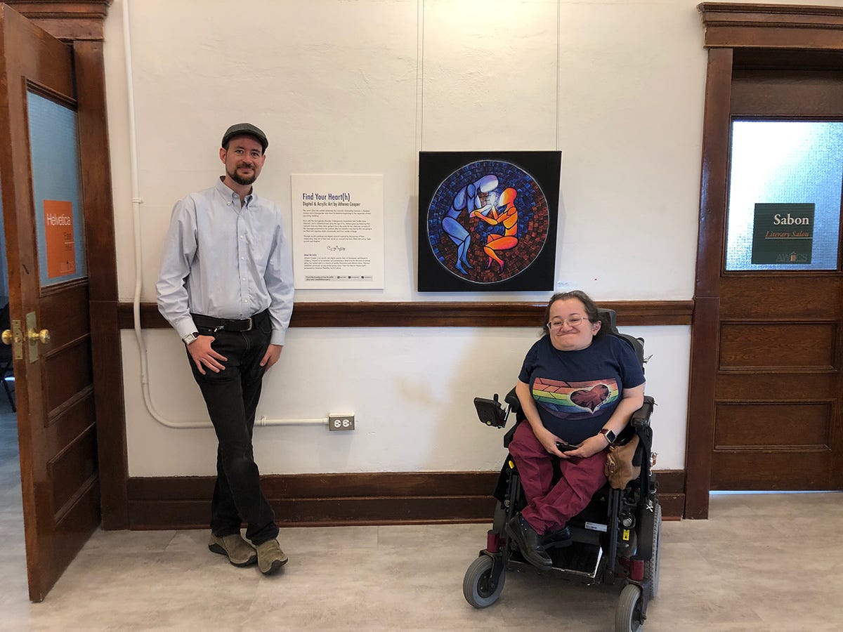 Stefan and Athena on either side of a large canvas print of the painting with the blue and orange figures. Stefan stands next to a large description panel with the title of the exhibit, "Find Your Heart(h)" and Athena sits in her power wheelchair next to the canvas print.
