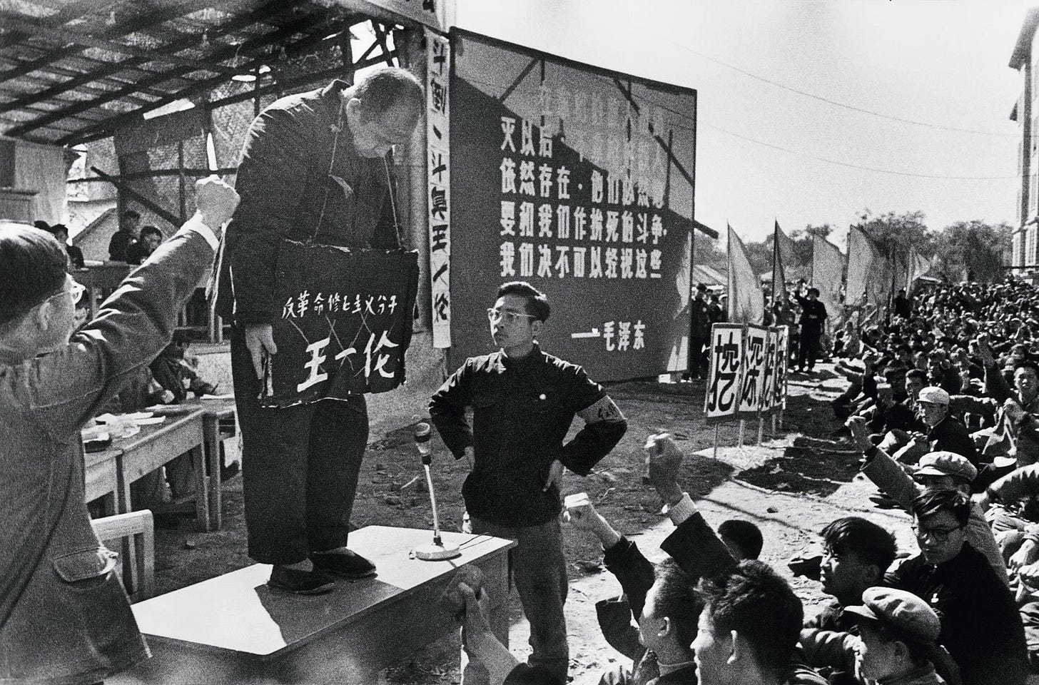 A man standing on a pedestal with his head hung in shame has a sign hung around his neck describing the offence he is accused of. A crowd of Red Guard and cultural revolutionaries sit around and watch as the man is shamed and berated into submission.