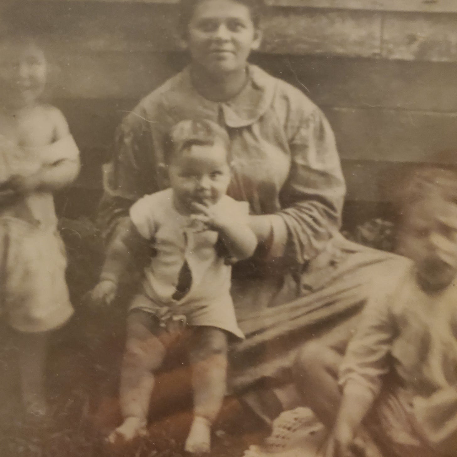 A black and white photo of a broad-faced woman and three small boys sitting in the dirt outside a clapboard house. 