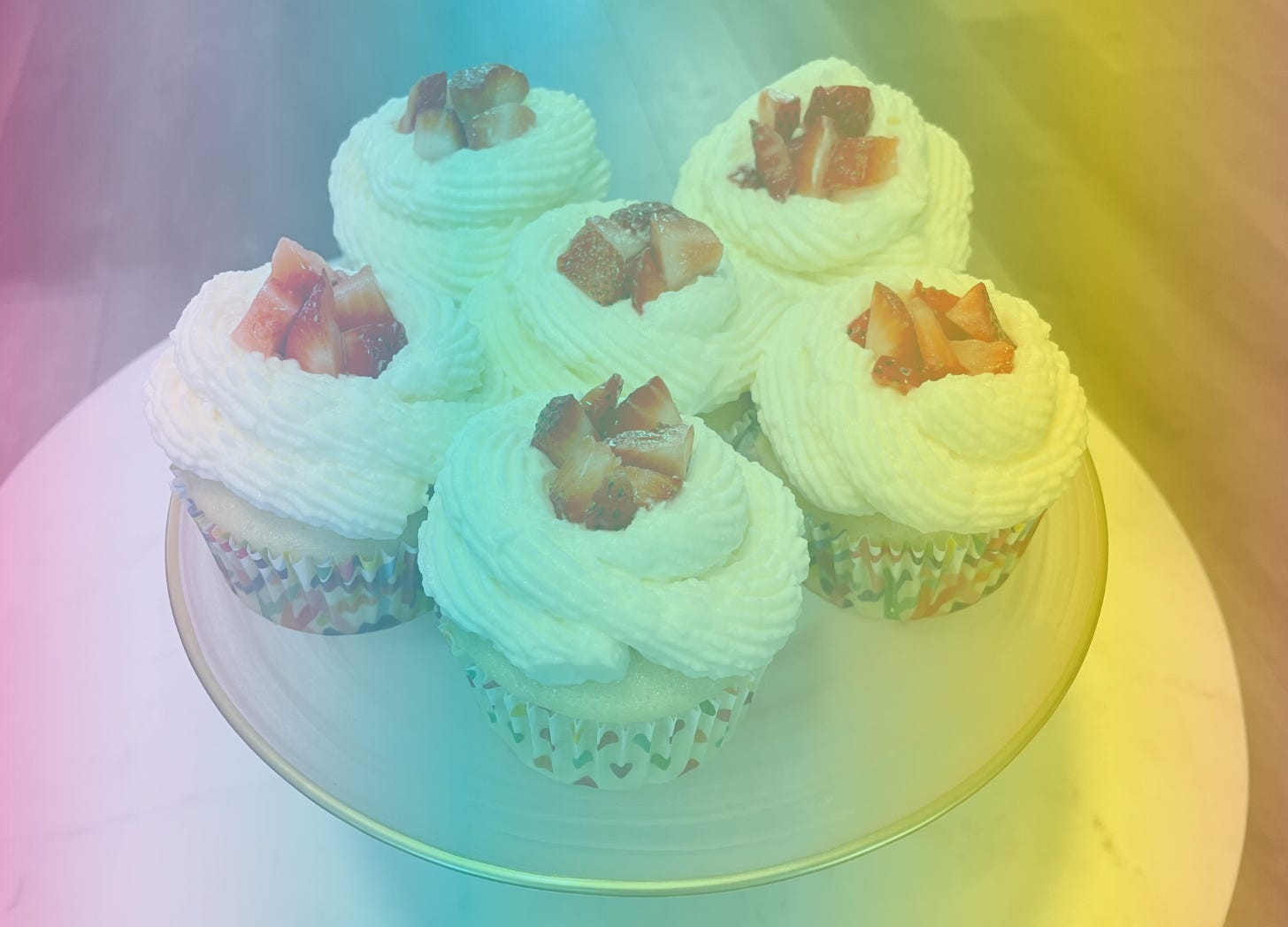 A rainbow-filtered photograph of 5 cupcakes on a plate, piled high with white whipped cream frosting and diced strawberries