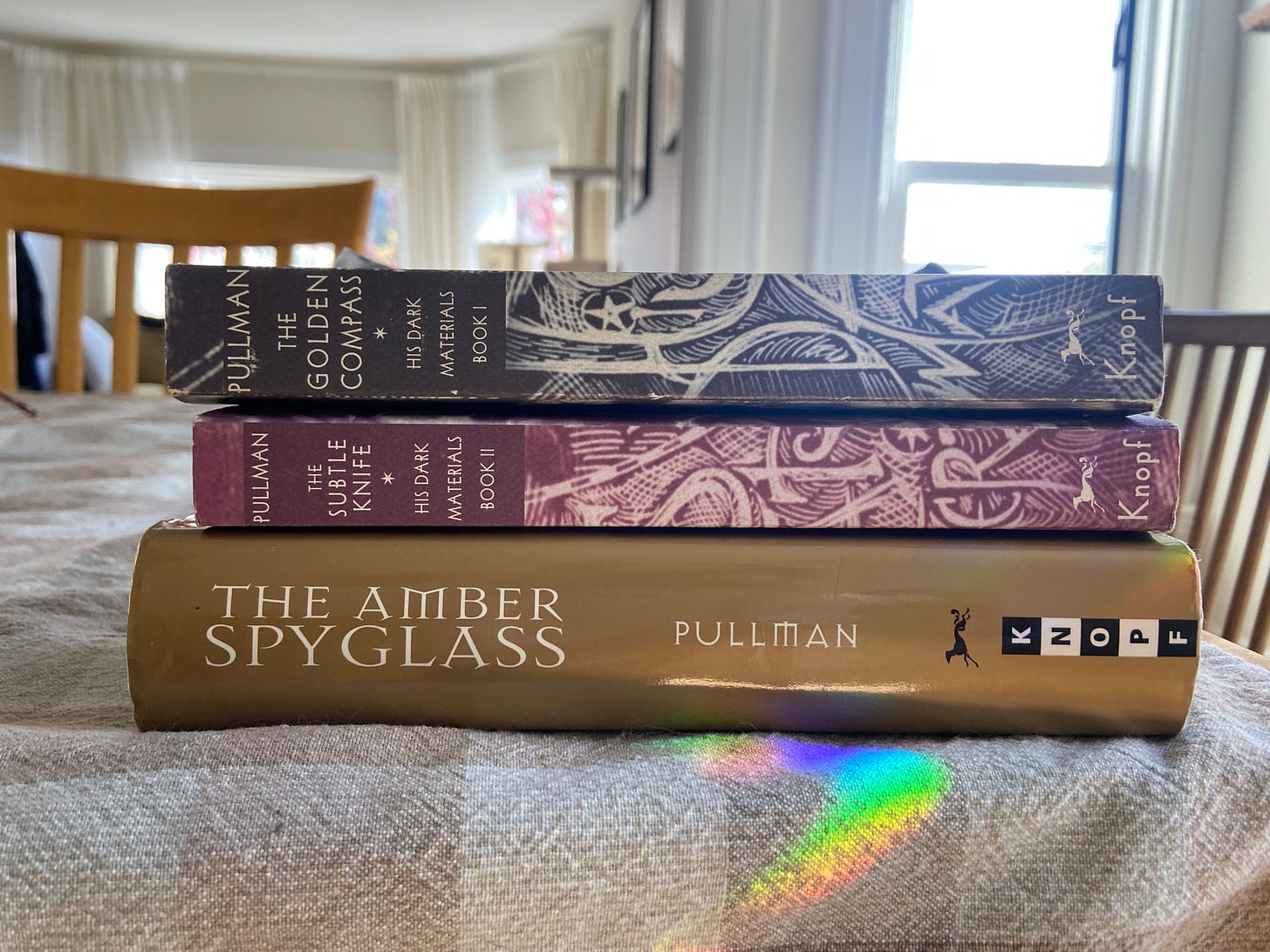 a photo of the three books in the His Dark Materials trilogy stacked on top of each other, on my dining room table. a rainbow from a lightcatcher rests on them. 