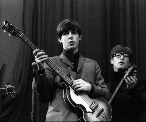 Paul McCartney holds his bass on stage while John Lennon tunes his guitar behind Paul