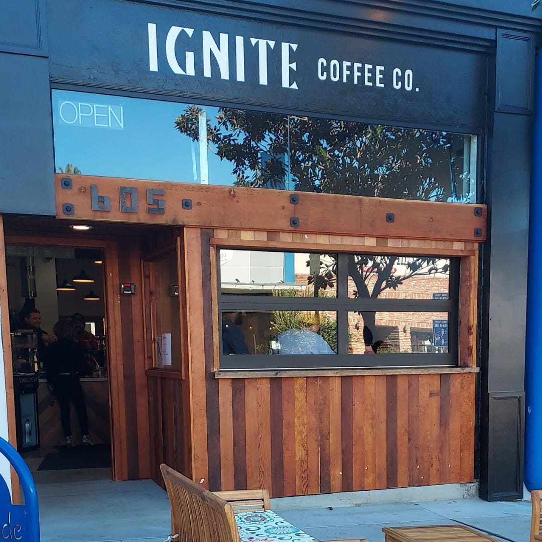 The entry of Ignite Coffee Company. The name is painted in white letters above a large window and the front door which is framed in reclaimed wood. A roll-up garage door style window to the right of the door is closed but coffee drinkers can be seen through the glass.
