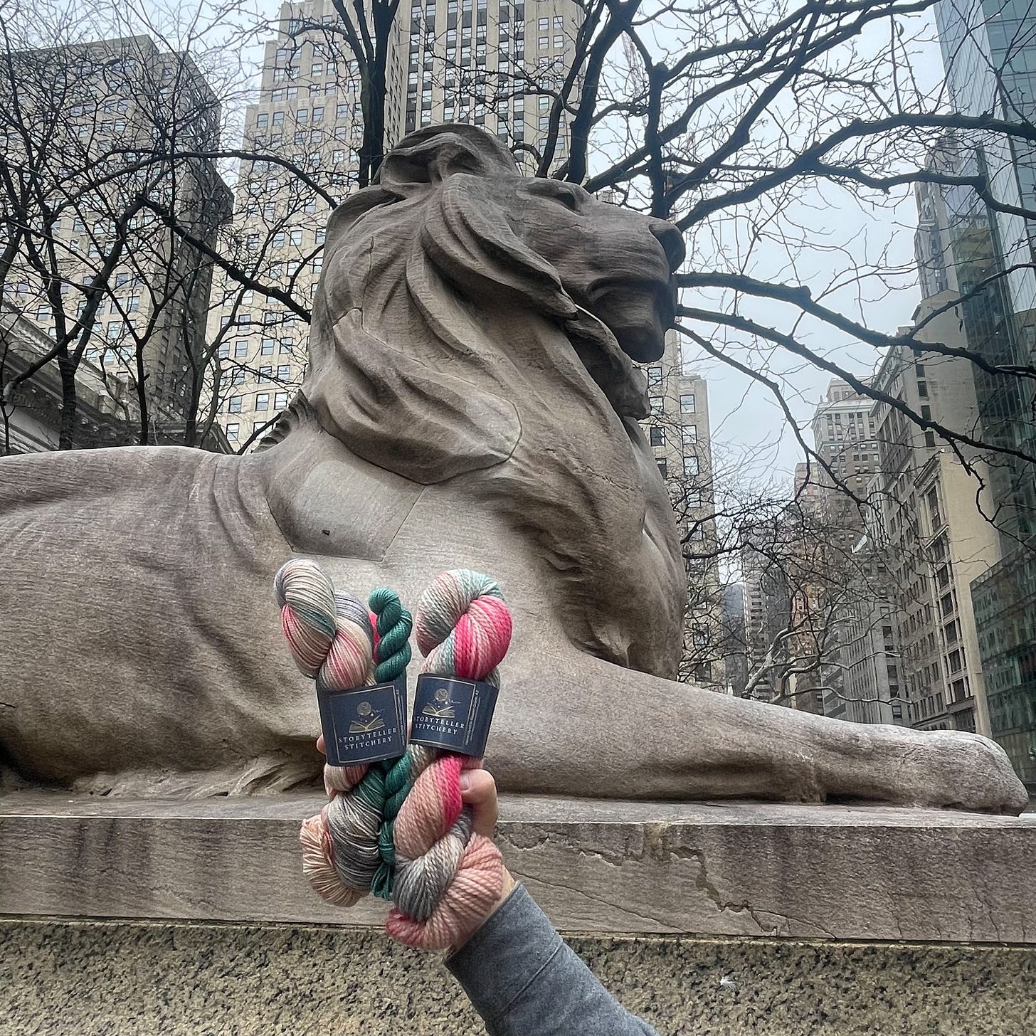Image of a lion statue, with New York buildings in the background and a hand holding up yarn. 