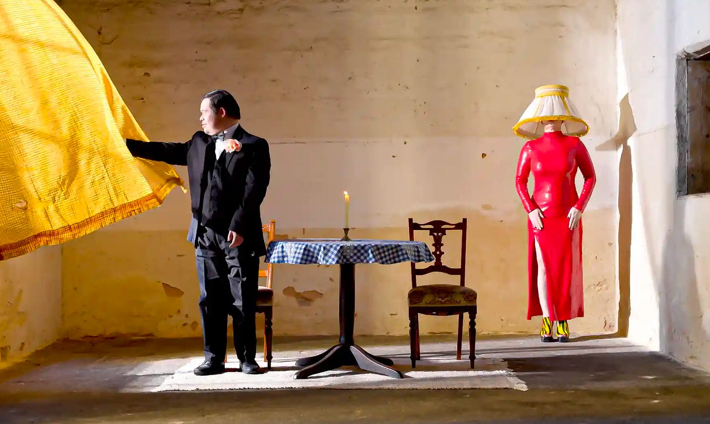 A masc actor in a three-piece suit is looking out a window covered by a yellow curtain, next to a table set for 2. In the back corner of the room/stage, a femme in a red latex suit is a floor lamp, with a shade covering her head.
