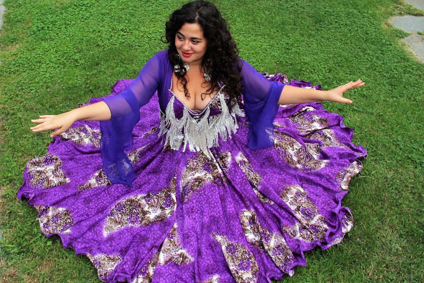 Ivana Nikolic è una donna con lunghi capelli castani e mossi, siede sul prato mentre indossa un ampio vestito viola con decorazioni grigie. Tiene le braccia aperte come in un morbido passo di danza. Ivana Nikolic è una donna con lunghi capelli castani e mossi, siede sul prato mentre indossa un ampio vestito viola con decorazioni grigie. Tiene le braccia aperte come in un morbido passo di danza.