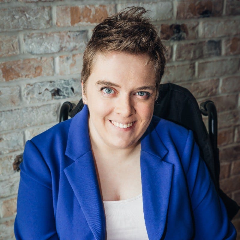 Lucy is a young woman wearing a blue jacket and white top.  She has blue eyes and a beaming smile.  her hair is short and brown in colour.  Lucy is sat in her wheelchair with a brick wall behind her.  