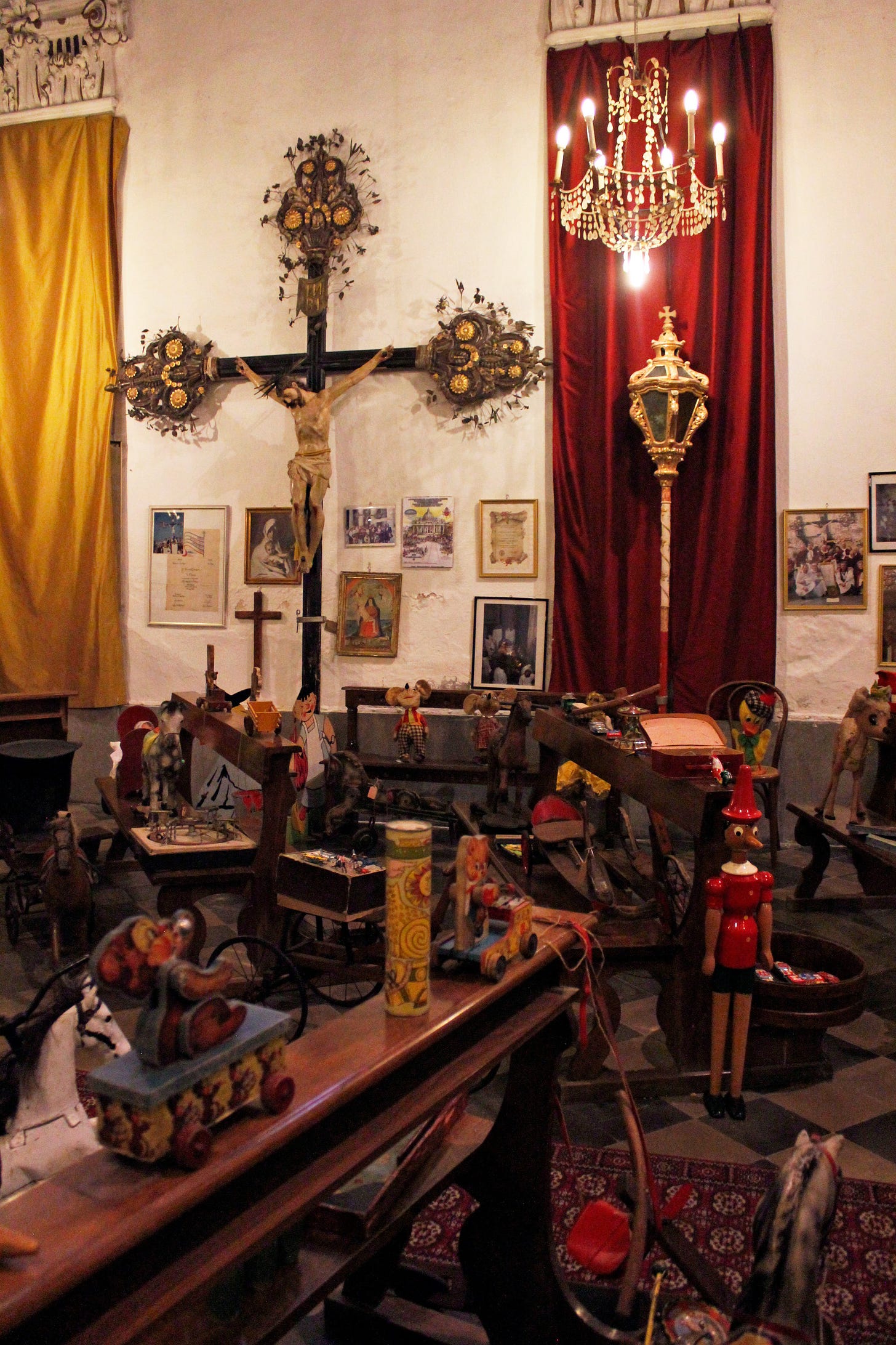 A photo taken in an oratory. There is a large crucifix on the wall and a chandelier overhead, but in the pews are dolls, wooden toys and stuffed animals.
