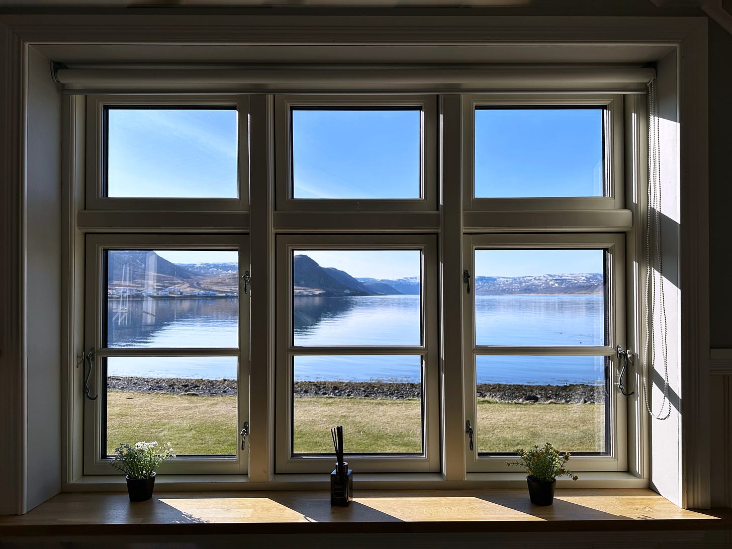 Morning light through three vertical windows that look over the fjord.  Blue skies and blue, calm water with spring green grass in front.