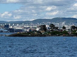 A view from a boat. Lots of deep-blue water, with a bit of land ahead. It's green and lush, with small cabins and houses dotted about. In the distance in the horizon is the mainland. Houses and buildings along the coast, and mountains in the background. The sky is blue with some white fluffy clouds.