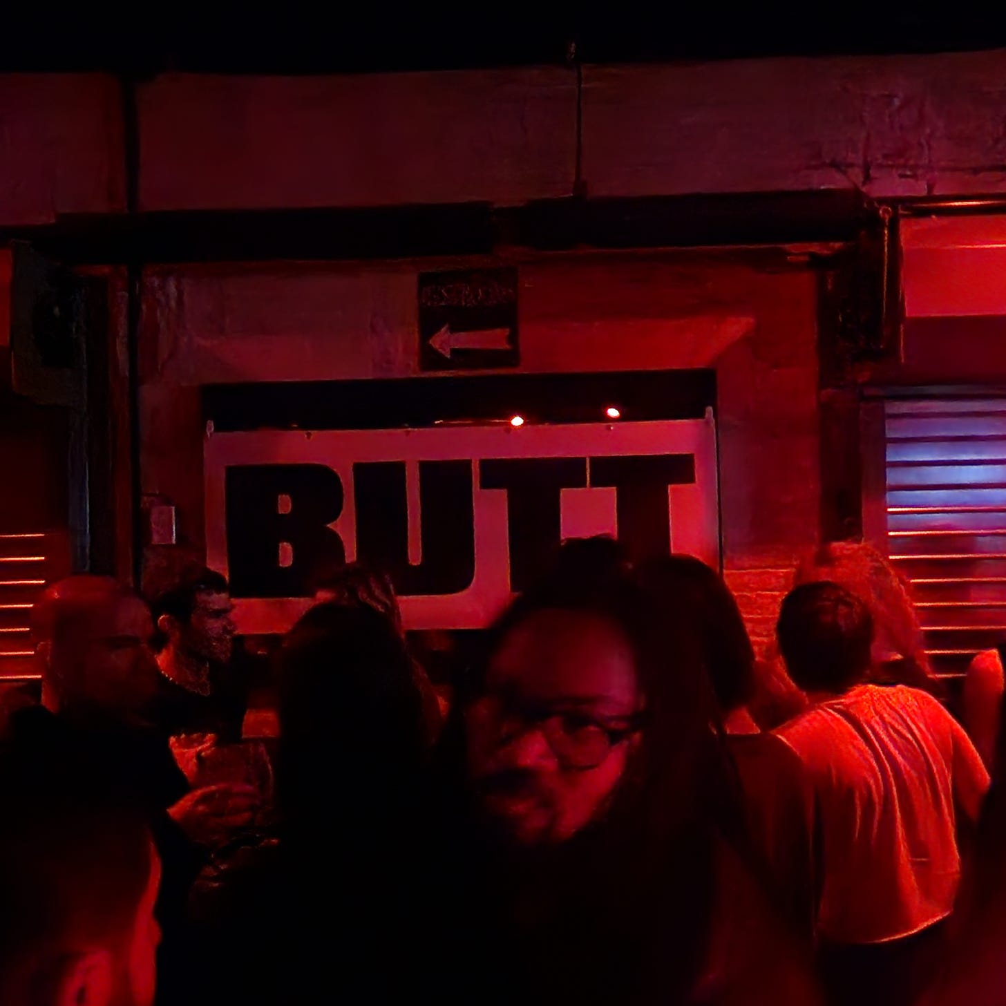 The image captures a crowded party atmosphere with warm red lighting, typical of a club setting. In the background, there's a prominent sign with the word 'BUTT' illuminated, indicating the theme or title of the event. Foreground shows silhouettes and partial views of attendees in casual party wear, engaged in conversation and socializing.