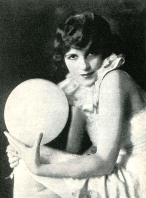 An old, black and white photo of a petite white, androgynous looking woman wearing a white short dress, her shoulders beared and a white ruff around her neck. She is holding a white globe.