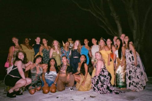 A photo taken on film of 21 femmes in long flowing dresses. The first row of femmes are crouching on their knees just above the sand of the beach they spent their last night on in Bali, Indonesia. The background is dark but you can still see the smiling faces and colorful prints of everyone;s dresses.