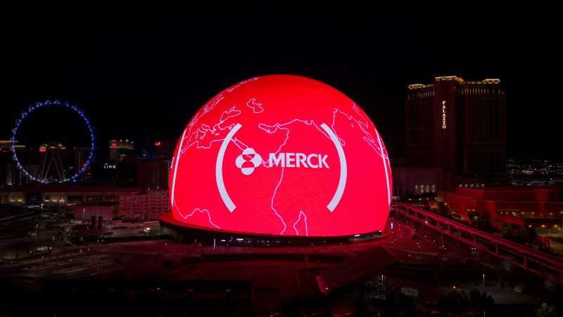 Grande redoma de mapa mundi vermelho com logo da Merck em cidade à noite.