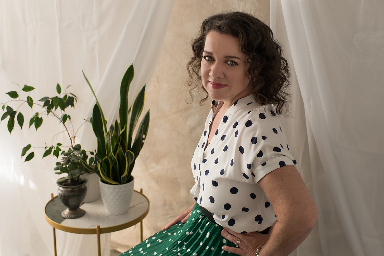 Tracy Byrne smiling at the camera and wearing a polka dot blouse and skirt with a table of potted plants beside her